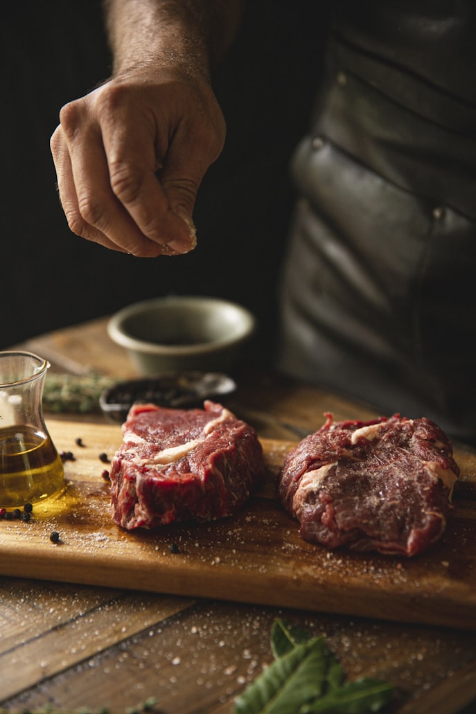 a person applying salt to raw steak on a cutting board